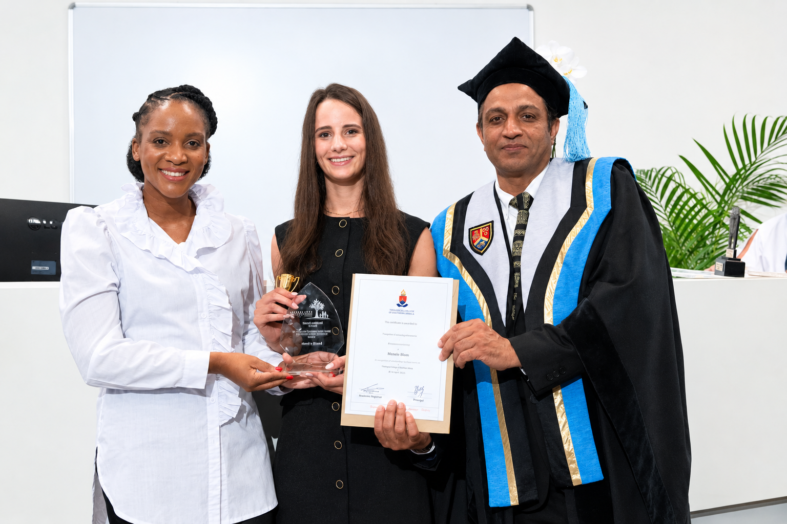 Three people at an award presentation, one holding a glass award and a framed certificate, beside a man in academic dress