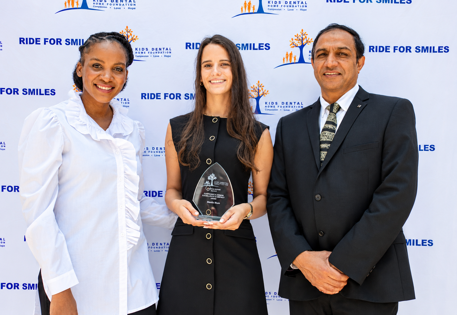 Three people at a Kids Dental Home Foundation Ride for Smiles event, one holding a glass award