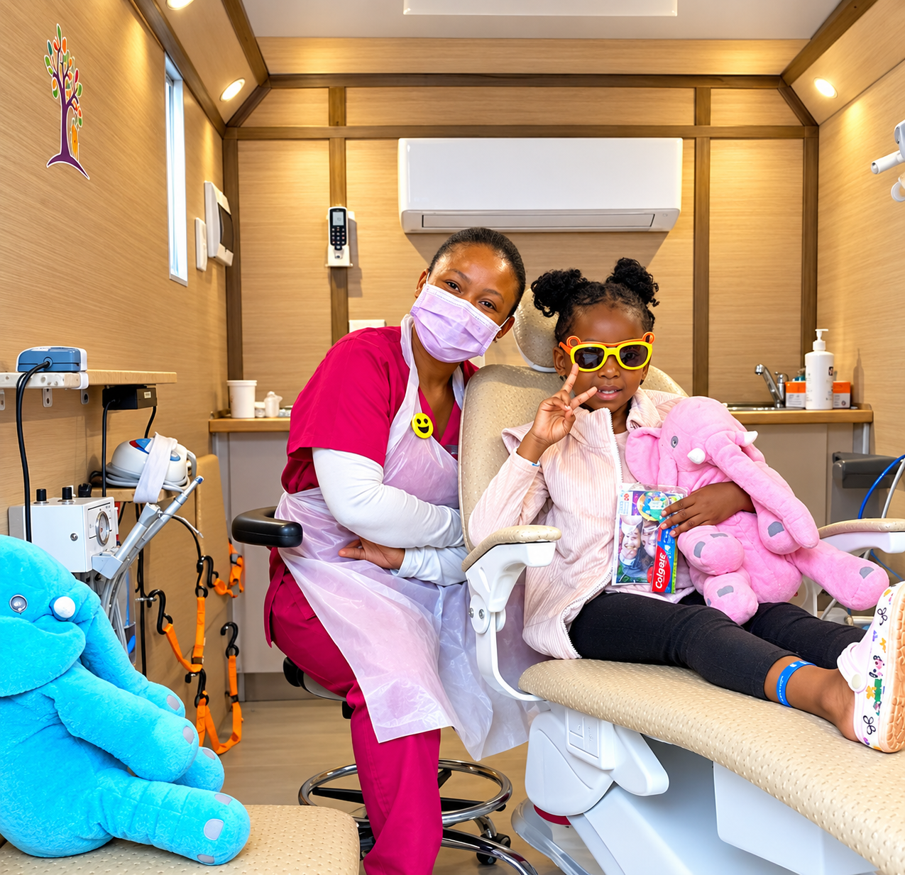 A girl in yellow sunglasses holding a pink toy elephant in a dental chair, making a peace sign beside a dental team member
