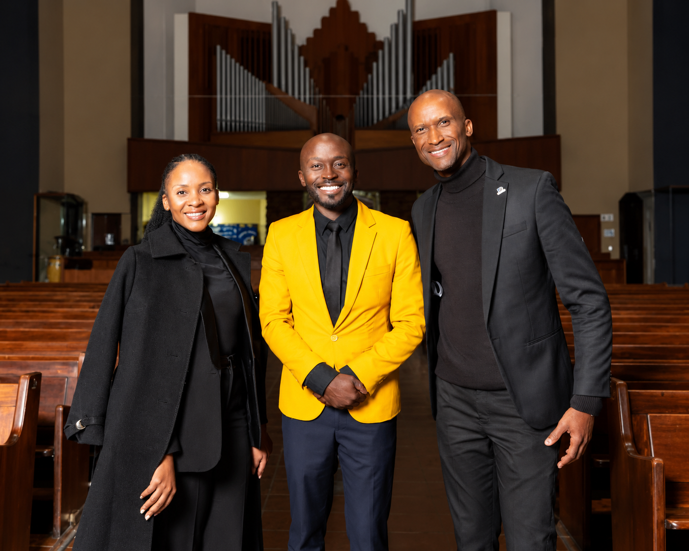 Three smiling people standing together in the aisle of a church, with the organ pipes behind them